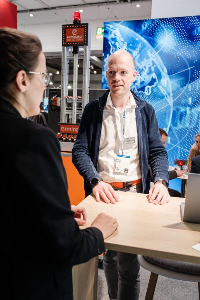 Gespräch am erminas-Messestand: erminas-Mitarbeiter am Stehtisch vor dem erminator Free Fall Tower und digitaler Weltkarten-Grafik.