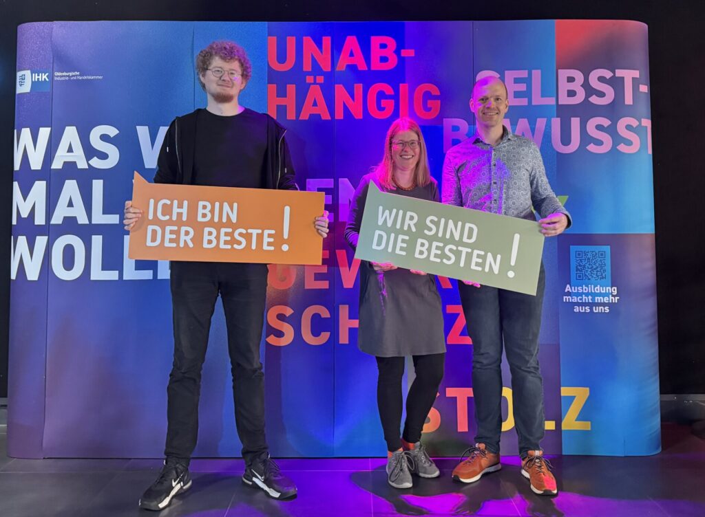 Drei Personen stehen vor einer bunten IHK-Fotowand mit der Aufschrift „Unabhängig“ und „Selbstbewusst“. Zwei von ihnen halten Schilder mit den Slogans „Ich bin der Beste!“ und „Wir sind die Besten!“. Die Szene ist in einem Innenraum mit Bühnenbeleuchtung in Blau- und Lilatönen aufgenommen.