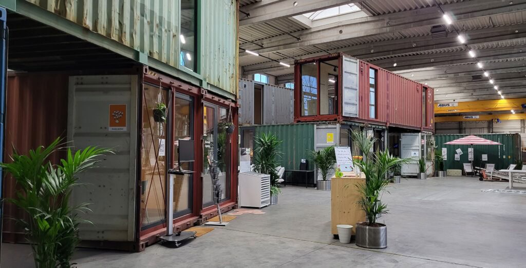 Zu Büro- und Arbeitsräumen umgebaute, gestapelte Schiffscontainer mit Pflanzen und Sitzbereichen in einer Industriehalle.